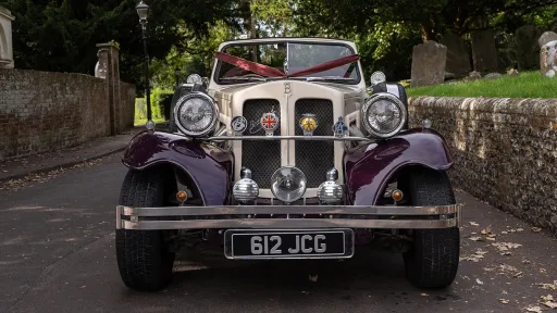 Front view of burgundy and cream four door Beauford convertible with chrome headlamps, wedding ribbons and folded burgundy roof outside stone church.