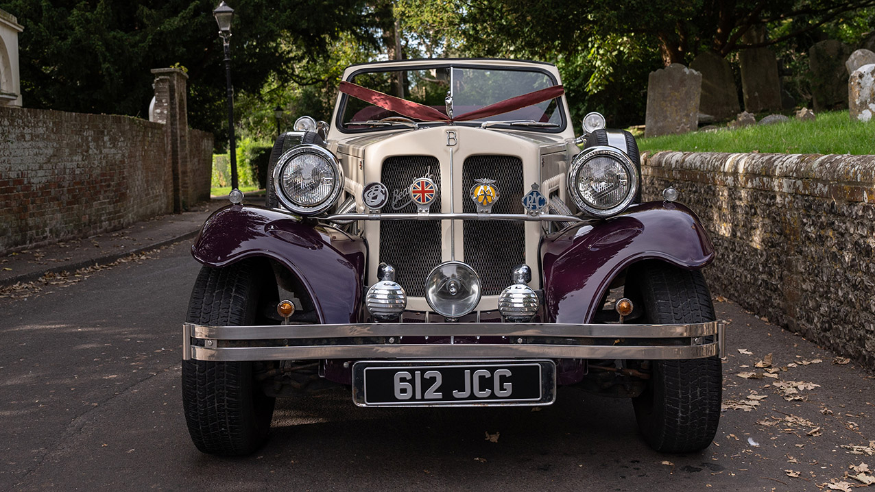 Front view of burgundy and cream four door Beauford convertible with chrome headlamps, wedding ribbons and folded burgundy roof outside stone church.