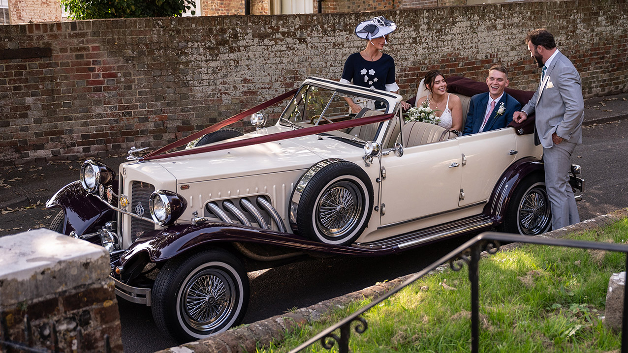 Bride and wedding guests beside burgundy and cream Beauford convertible with roof folded down crossing stone bridge.