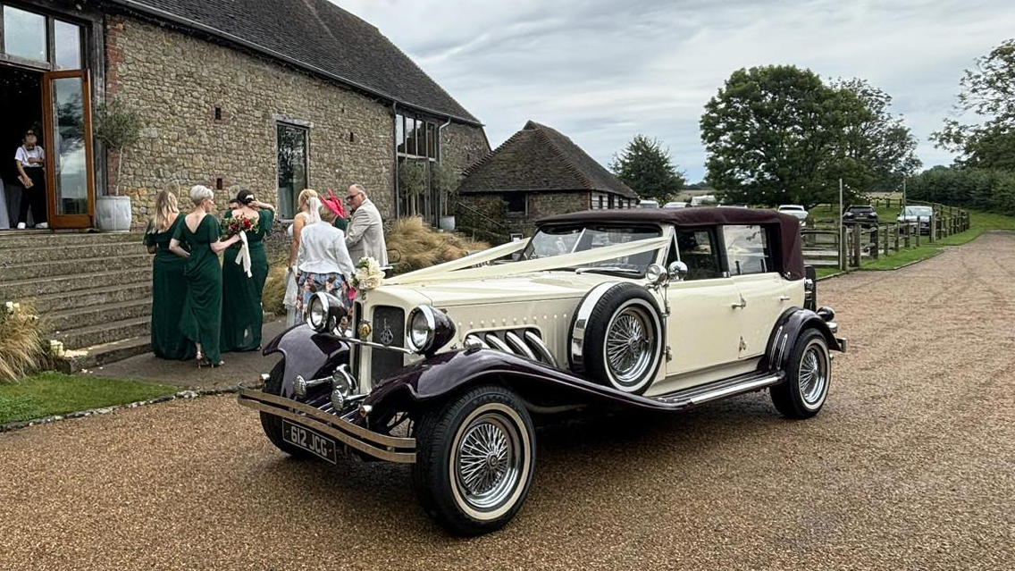 Burgundy and cream Beauford convertible with burgundy soft top roof raised outside country wedding venue.