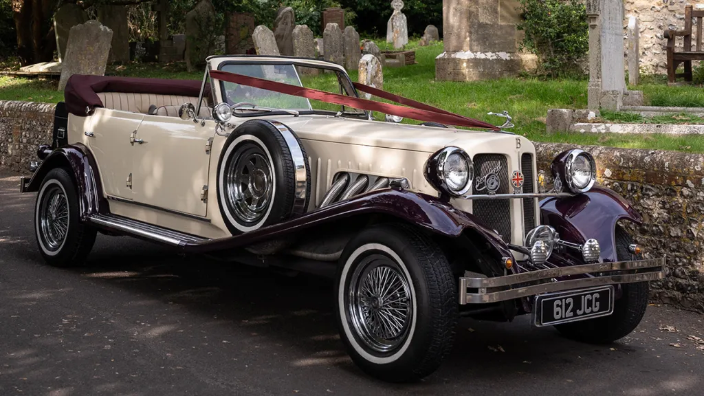 Side view of burgundy and cream Beauford convertible with open roof parked beside churchyard wall.