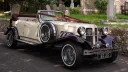 Side view of burgundy and cream Beauford convertible with open roof parked beside churchyard wall.