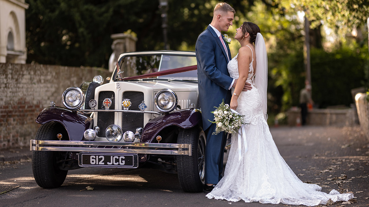 Bride and groom standing beside burgundy and cream Beauford convertible outside historic church in Wiltshire.