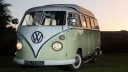 Front view of cream and green Volkswagen Split Screen campervan parked at sunset with headlights illuminated.
