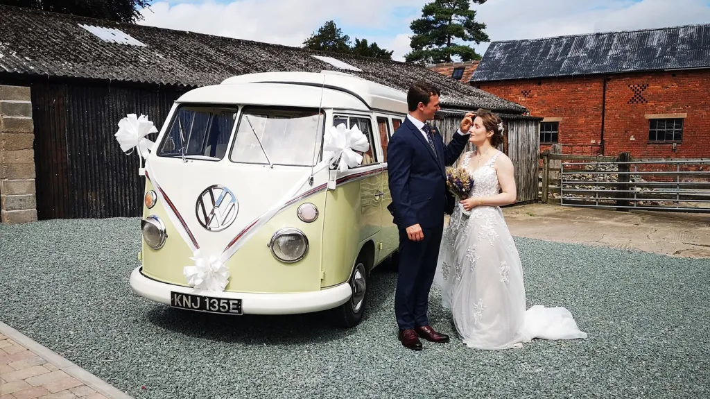 Bride and groom standing beside cream and green Volkswagen Split Screen campervan at rustic wedding venue.