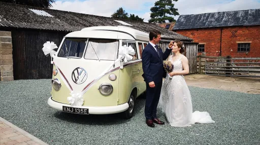 Bride and groom standing beside cream and green Volkswagen Split Screen campervan at rustic wedding venue.