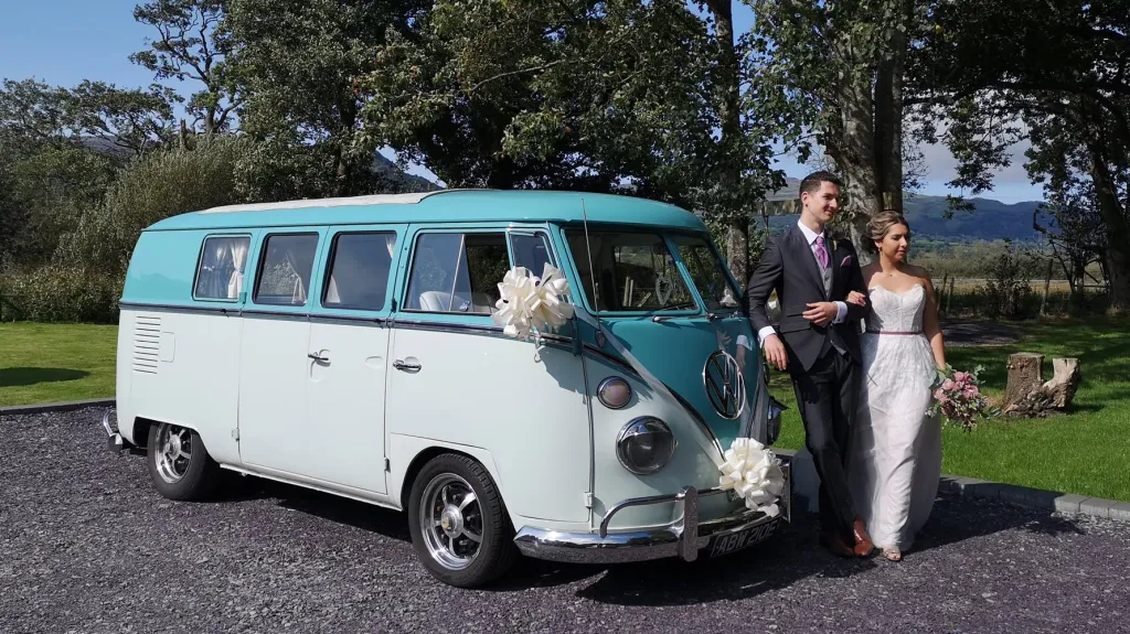 Bride and groom standing beside turquoise and white 1967 Volkswagen Split Screen campervan at wedding venue.
