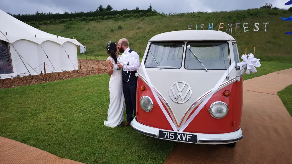 Bride and groom standing beside red and white 1962 Volkswagen Split Screen Microbus at outdoor wedding venue near Wrexham.