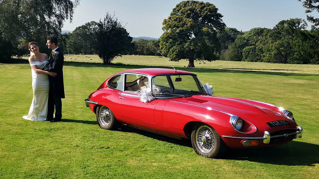 Bride and groom standing beside red 1969 Jaguar E Type parked on lawn at country wedding venue.