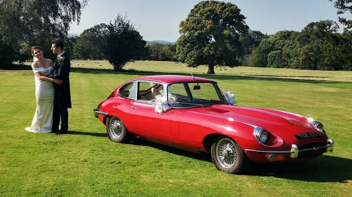 Bride and groom standing beside red 1969 Jaguar E Type parked on lawn at country wedding venue.