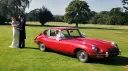 Bride and groom standing beside red 1969 Jaguar E Type parked on lawn at country wedding venue.