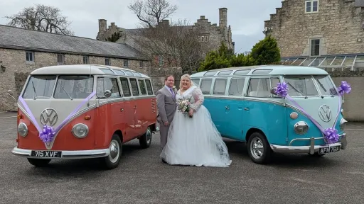 Bride and groom standing between two decorated Volkswagen Split Screen campervans at wedding venue.