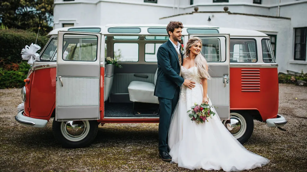 Bride and groom beside red and white Volkswagen Split Screen 23 Window Microbus with side doors open.