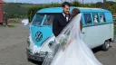 Bride and groom standing beside turquoise and white 1967 Volkswagen Split Screen campervan decorated with wedding ribbons.