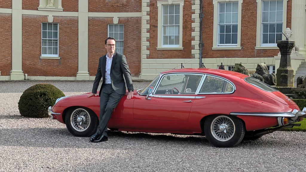Full left side profile of red 1969 Jaguar E Type coupe parked outside country house venue.