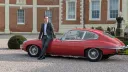 Full left side profile of red 1969 Jaguar E Type coupe parked outside country house venue.