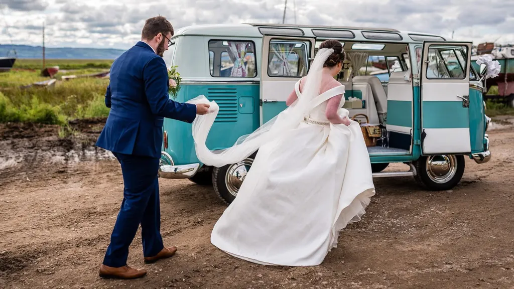Groom helping the bride into a blue and white Volkswagen Split Screen Samba campervan.