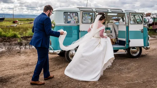 Groom helping the bride into a blue and white Volkswagen Split Screen Samba campervan.
