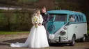 Newly married couple posing beside turquoise and white Volkswagen Split Screen campervan at countryside wedding venue.