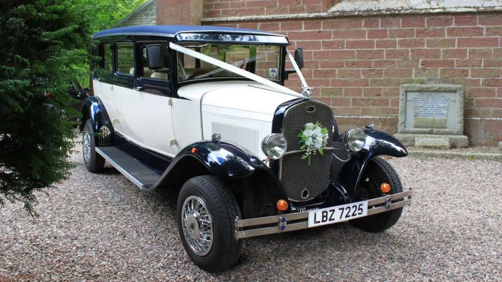 Front three quarter view of navy blue and ivory Bramwith limousine with chrome radiator, side mounted spare wheel and wedding ribbons.