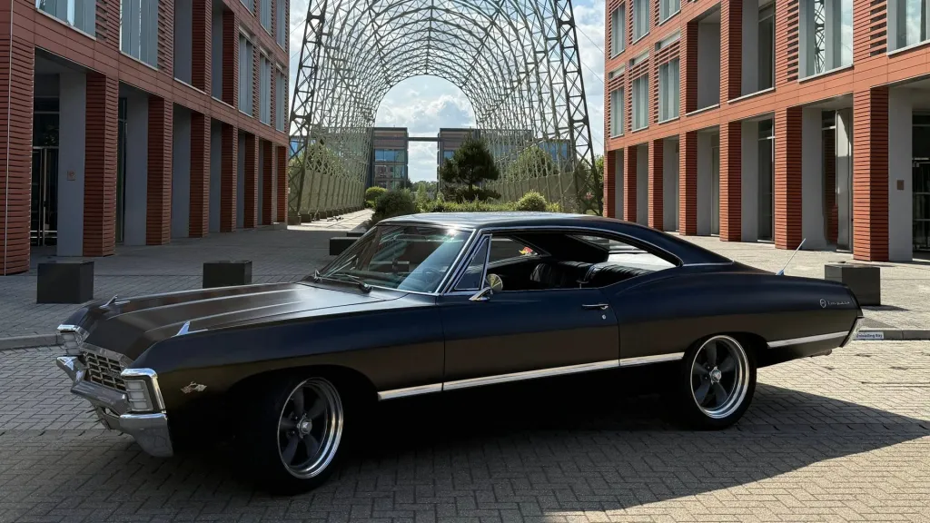 Side view of black 1967 Chevrolet Impala Fastback parked outside modern glass building.