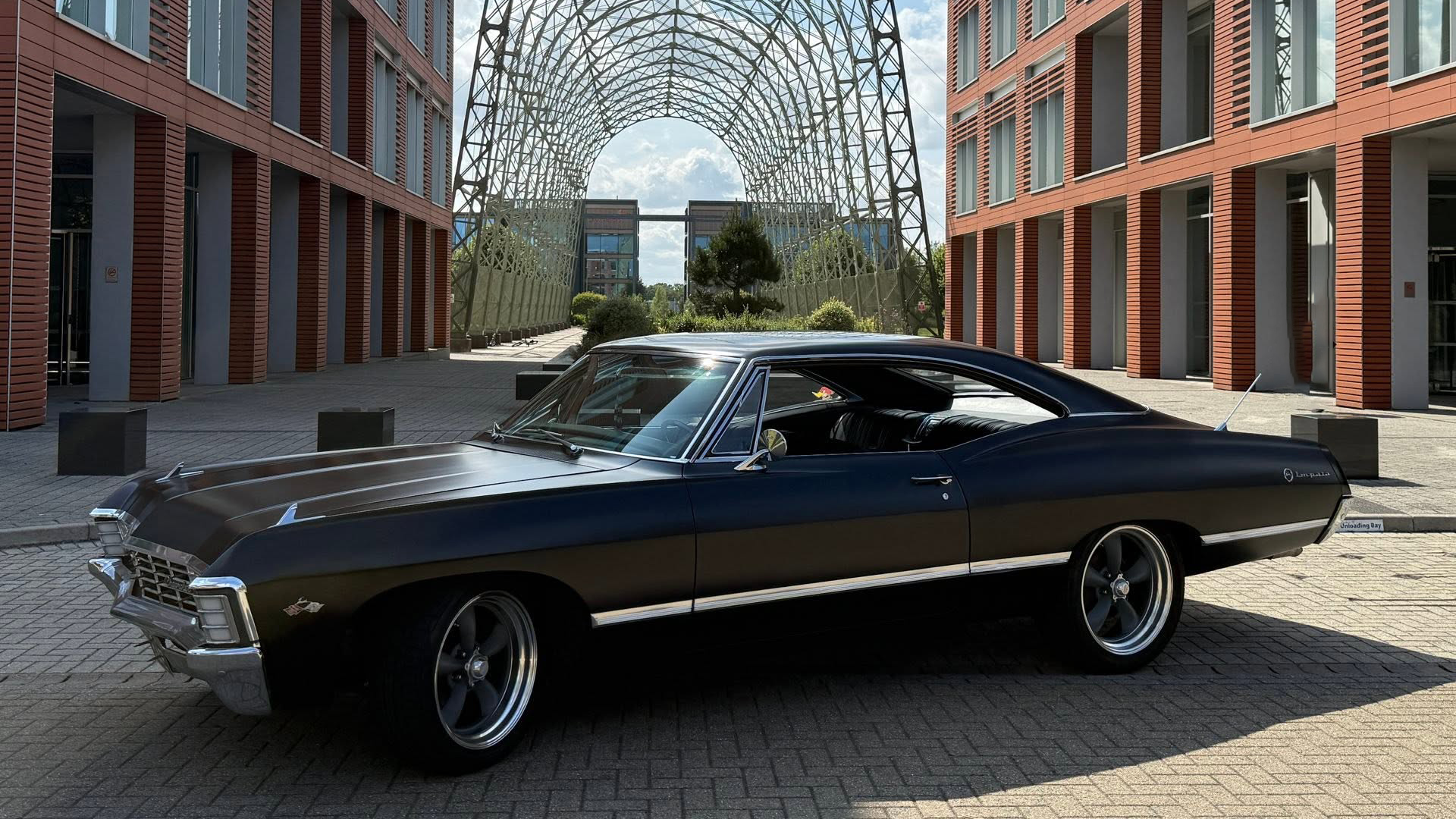 Side view of black 1967 Chevrolet Impala Fastback parked outside modern glass building.
