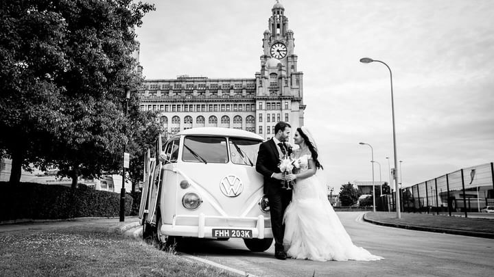 Black and white photograph of bride and groom beside cream 1972 Volkswagen Split Screen campervan outside cathedral.