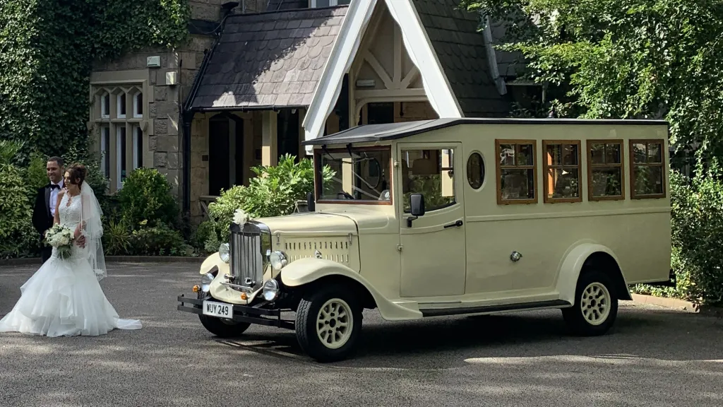 Bride standing beside Old English White Asquith vintage wedding bus outside countryside church.