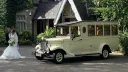 Bride standing beside Old English White Asquith vintage wedding bus outside countryside church.
