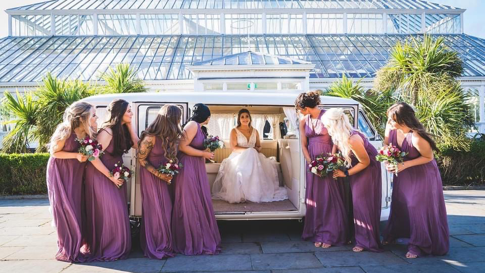 Bride and bridesmaids gathered around cream 1972 Volkswagen Split Screen campervan outside wedding venue.
