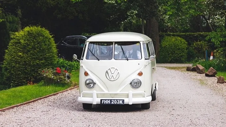 Front view of cream 1972 Volkswagen Split Screen campervan decorated with wedding ribbons entering wedding venue.