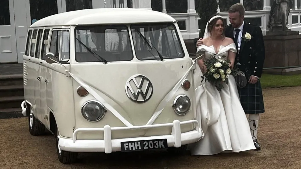 Bride and groom beside cream 1972 Volkswagen Split Screen campervan decorated with white wedding ribbons.