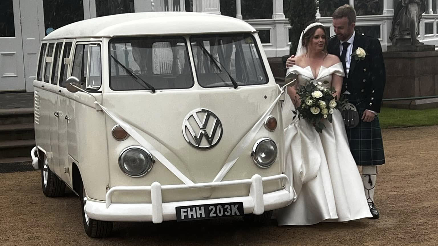Bride and groom beside cream 1972 Volkswagen Split Screen campervan decorated with white wedding ribbons.