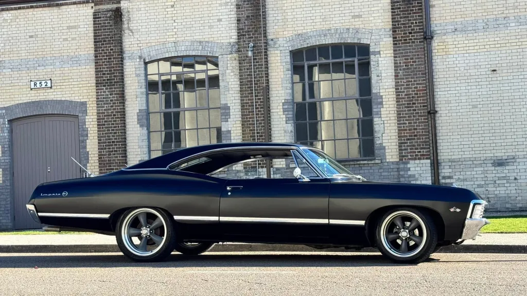 Right side view of black 1967 Chevrolet Impala Fastback parked outside industrial heritage building.