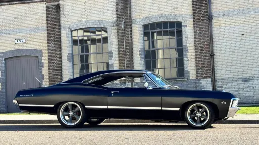 Right side view of black 1967 Chevrolet Impala Fastback parked outside industrial heritage building.