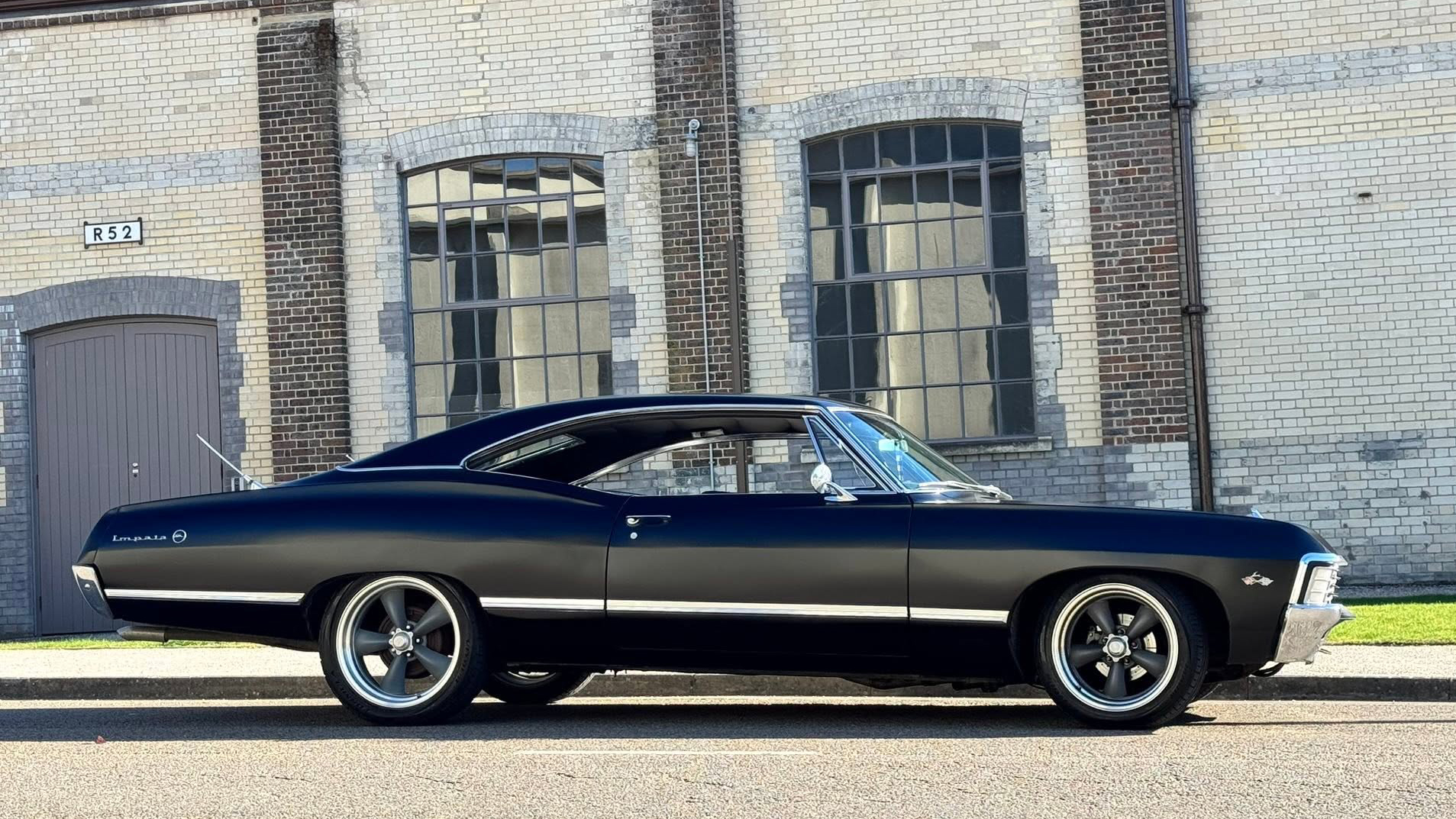 Right side view of black 1967 Chevrolet Impala Fastback parked outside industrial heritage building.