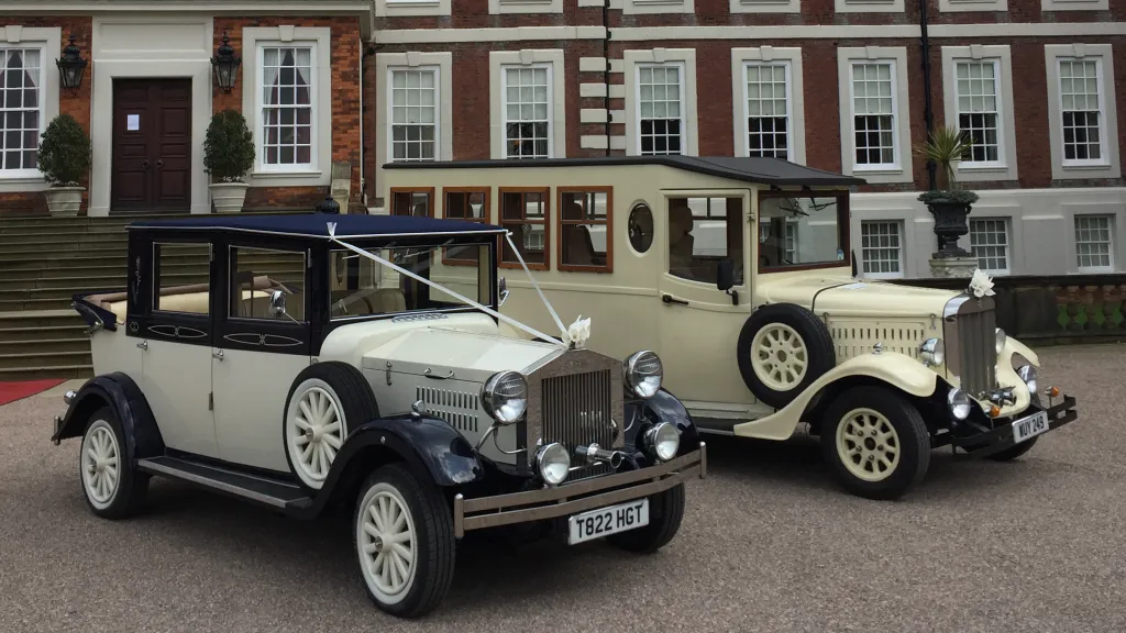 Old English White Asquith vintage wedding bus parked beside matching vintage wedding car outside hotel entrance.