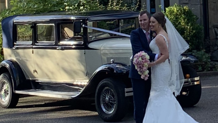 Bride and groom beside navy blue and ivory Bramwith Landaulette decorated with white wedding ribbons.