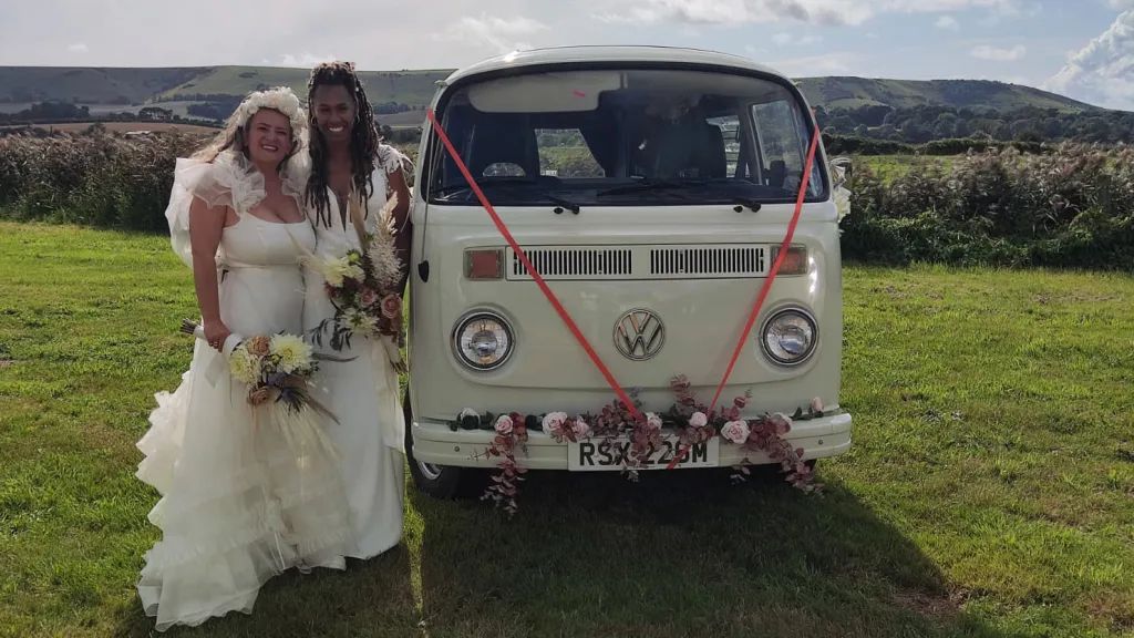 Two brides standing beside white 1974 Volkswagen Bay Window campervan decorated with wedding flowers in the Essex countryside.