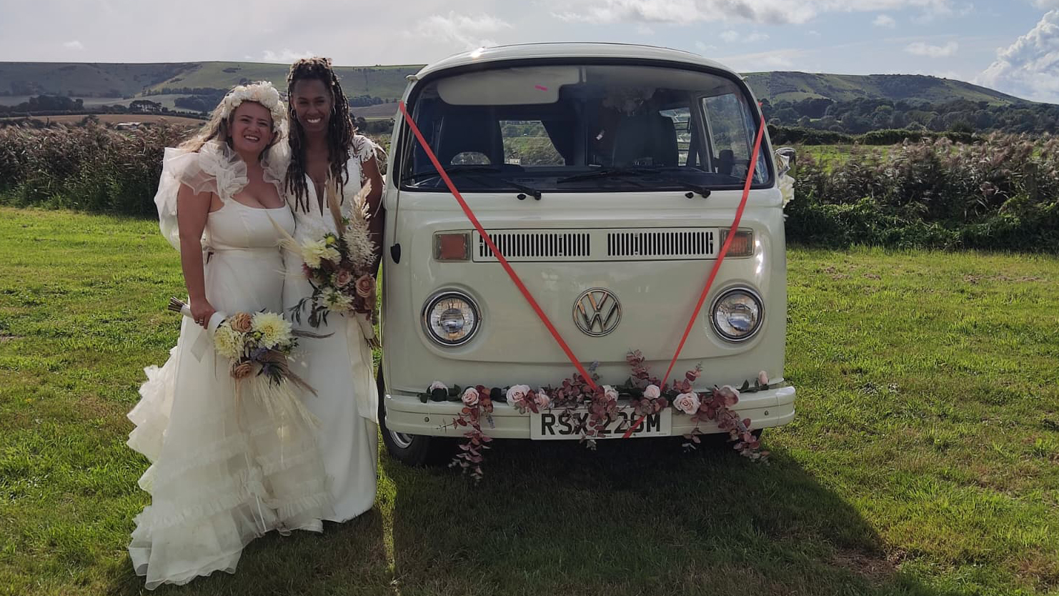 Two brides standing beside white 1974 Volkswagen Bay Window campervan decorated with wedding flowers in the Essex countryside.