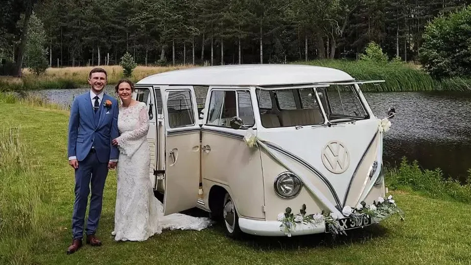 Bride and groom standing beside cream and beige 1964 VW Split Screen Campervan decorated with wedding flowers in Essex.