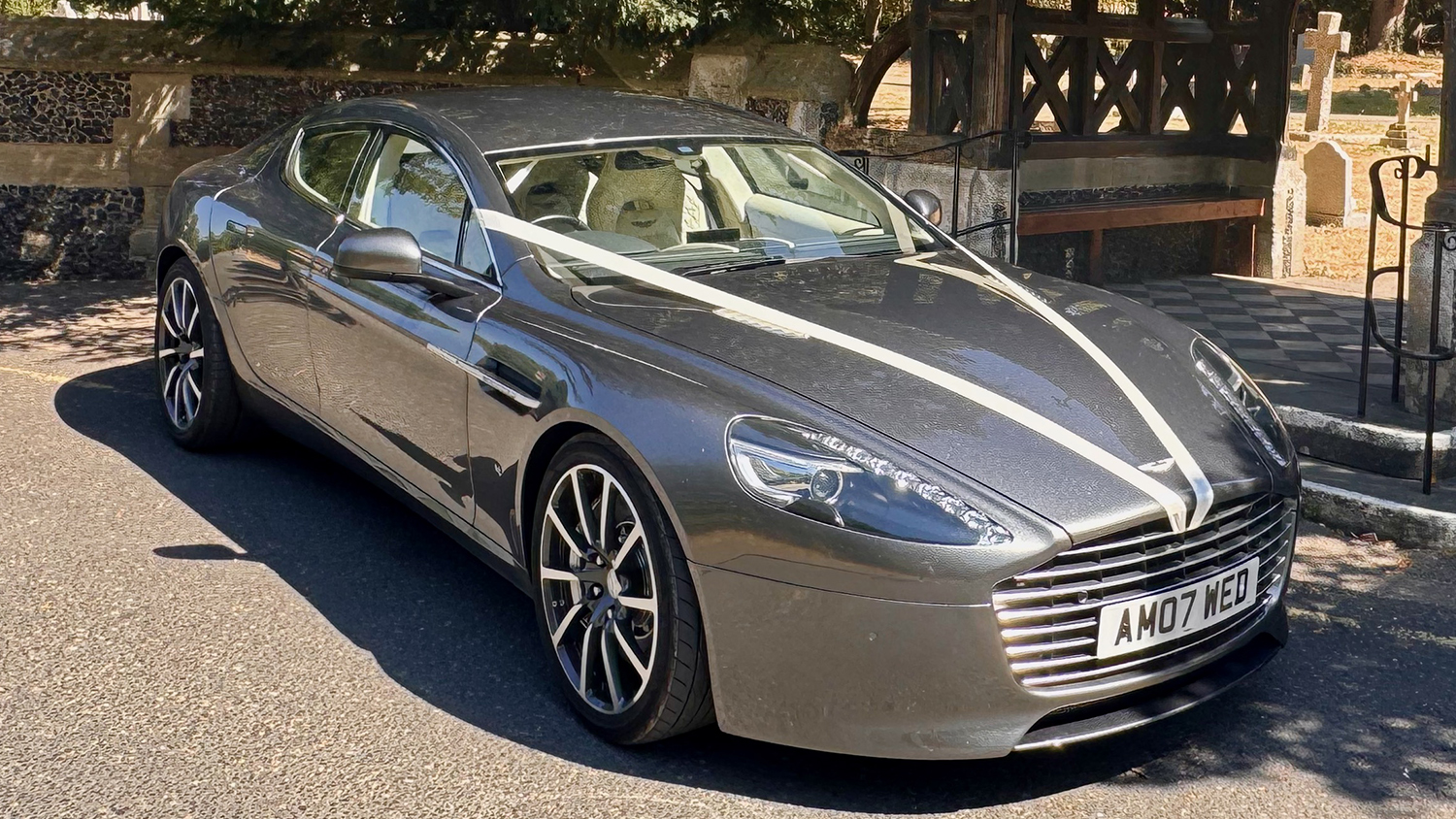 Front three quarter view of metallic silver Aston Martin Rapide S V12 with chrome grille and wedding ribbons.
