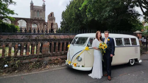 Groom with her bride holding a yellow sunflower standing in front of a classic campervan dressed with matching yellow ribbons and bows parked outside a church in Essex