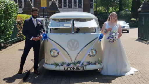 Bride and groom standing on either side of a cream classic campervan dressed with royal blue bows and white ribbons and matching white and blue garland of flowers on the bumper