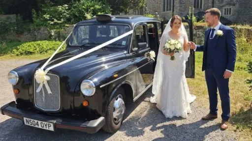 Bride and groom standing beside a classic black London-style taxi decorated with ivory wedding ribbons in Barry, South Wales.