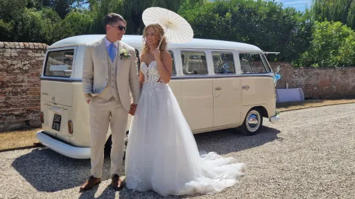 Groom in a cream suit holding hand with his Bride wearing a white dress and holding a White Umbrella with a cream and white classic wedding camperavn in the background