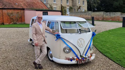 Chauffeur wearing a cream suit standing beside the cream splitscreen campervan with Royal Blue Ribbons and Bows