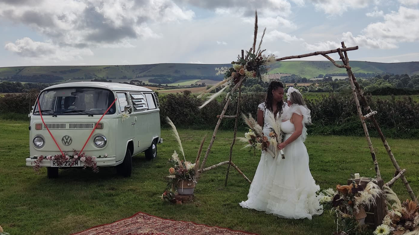 Two Brides wearing white dresses posing for phtoos with a classic white VW Campervan in the background