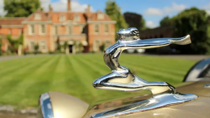 Close-up of chrome Beauford bonnet mascot on gold vintage-style wedding car with a Wiltshire country house in the background.