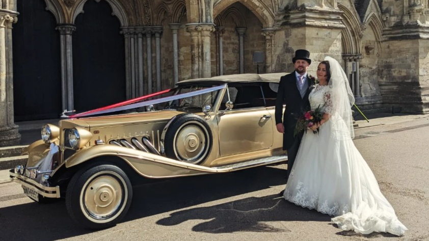 Bride and groom standing beside gold Beauford Convertible wedding car with black soft-top roof folded down.